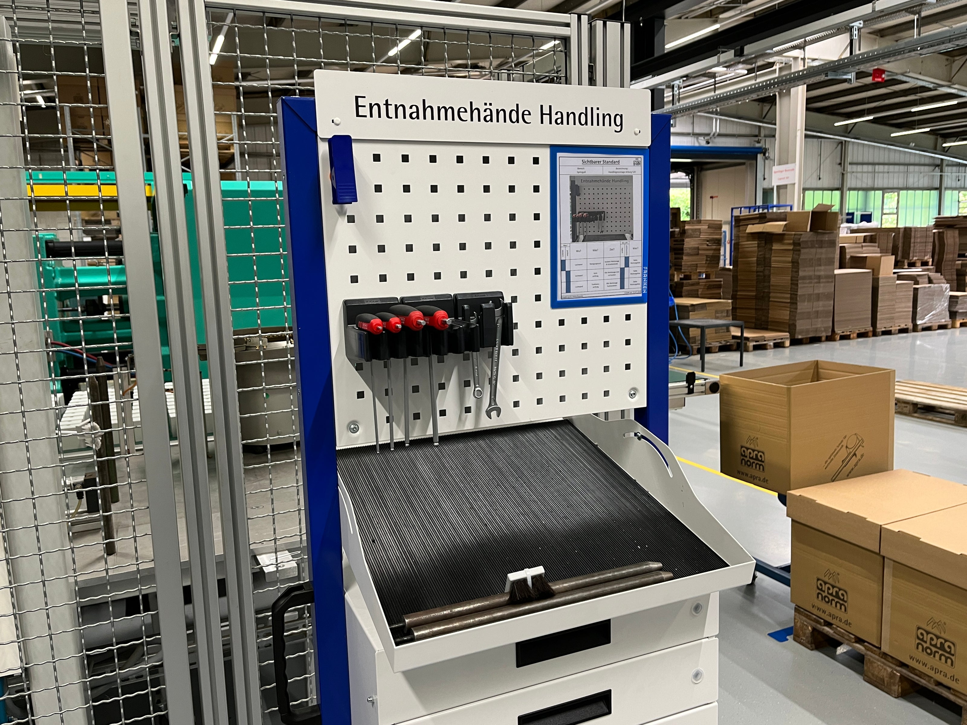 Prozessinsel zur Entnahmehandhabung in einer Produktionsumgebung. Die Station verfügt über ein Rasterblech mit befestigten Werkzeugen wie Schraubendrehern und einem Schraubenschlüssel, sowie eine geneigte Arbeitsfläche mit rutschfester Oberfläche. Im unteren Bereich befinden sich Schubladen zur Werkzeugaufbewahrung.